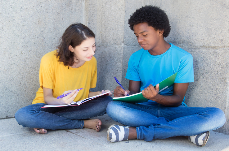 two students sitting down on a concrete floor and leaning against a concrete wall and sharing class notes