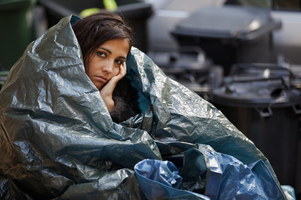 A young woman in the cold sad and covered by a plastic bag while sitting on the street pvaement.