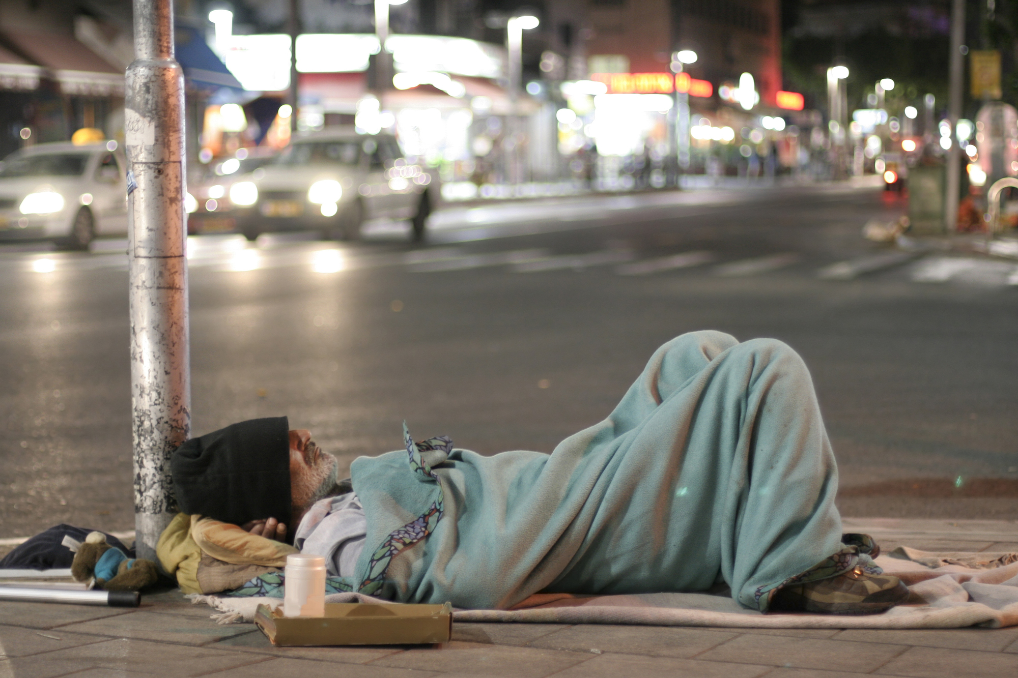 Homeless man is sleeping on the sidewalk of a busy avenue in the middle of the night, covered by a blanket.